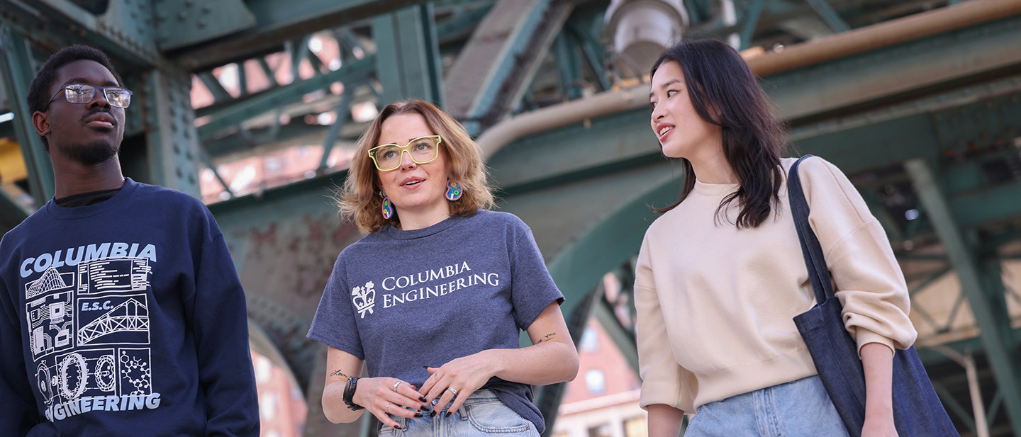 Three Columbia Engineering students walk together under a large bridge in New York City.